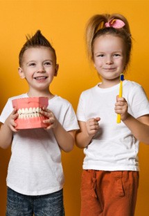 Three children dressed casually holding over-sized toothbrushes and a set of model teeth in front of goldenrod yellow background